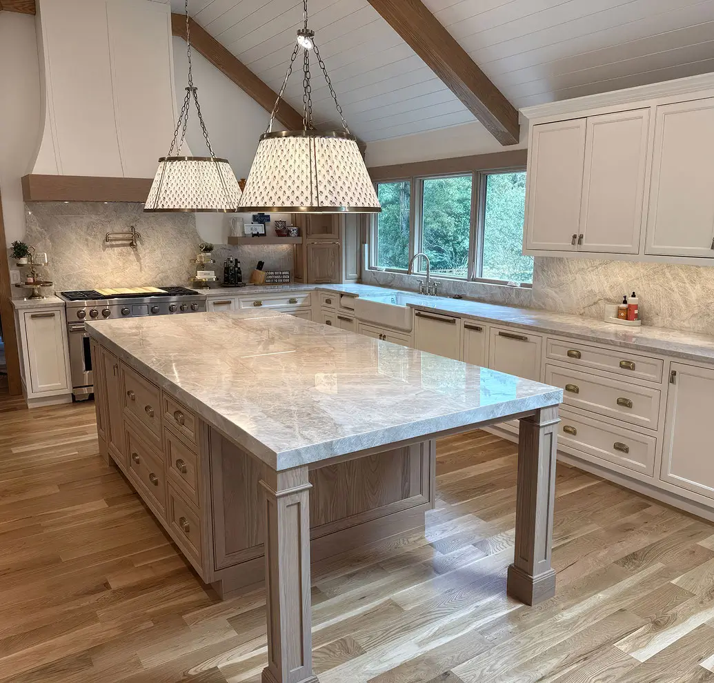 Bright kitchen with a large marble island, matching backsplash, white cabinets, wood beams, and pendant lights
